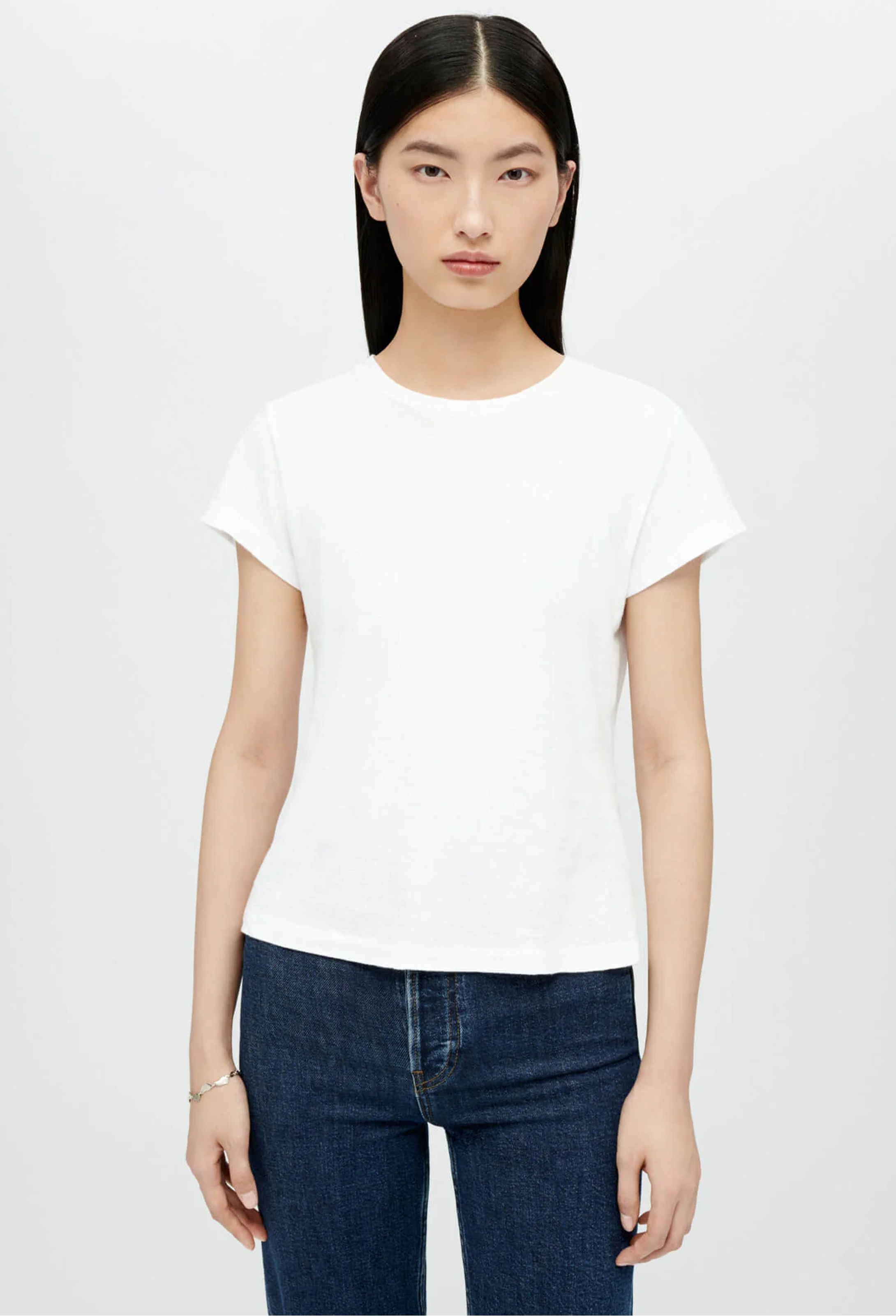 Model wears white crewneck t-shirt with dark wash blue jeans. She stands in front of a white background. 