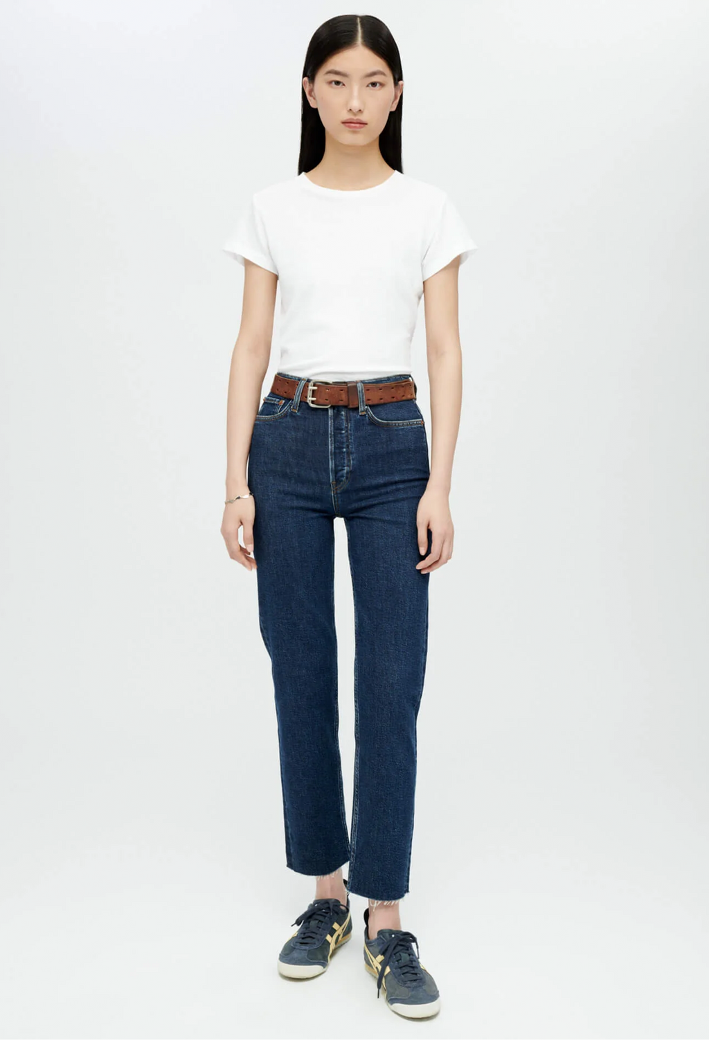 Model wears white crewneck t-shirt with dark wash blue jeans and brown belt and navy sneakers. She stands in front of a white background. 