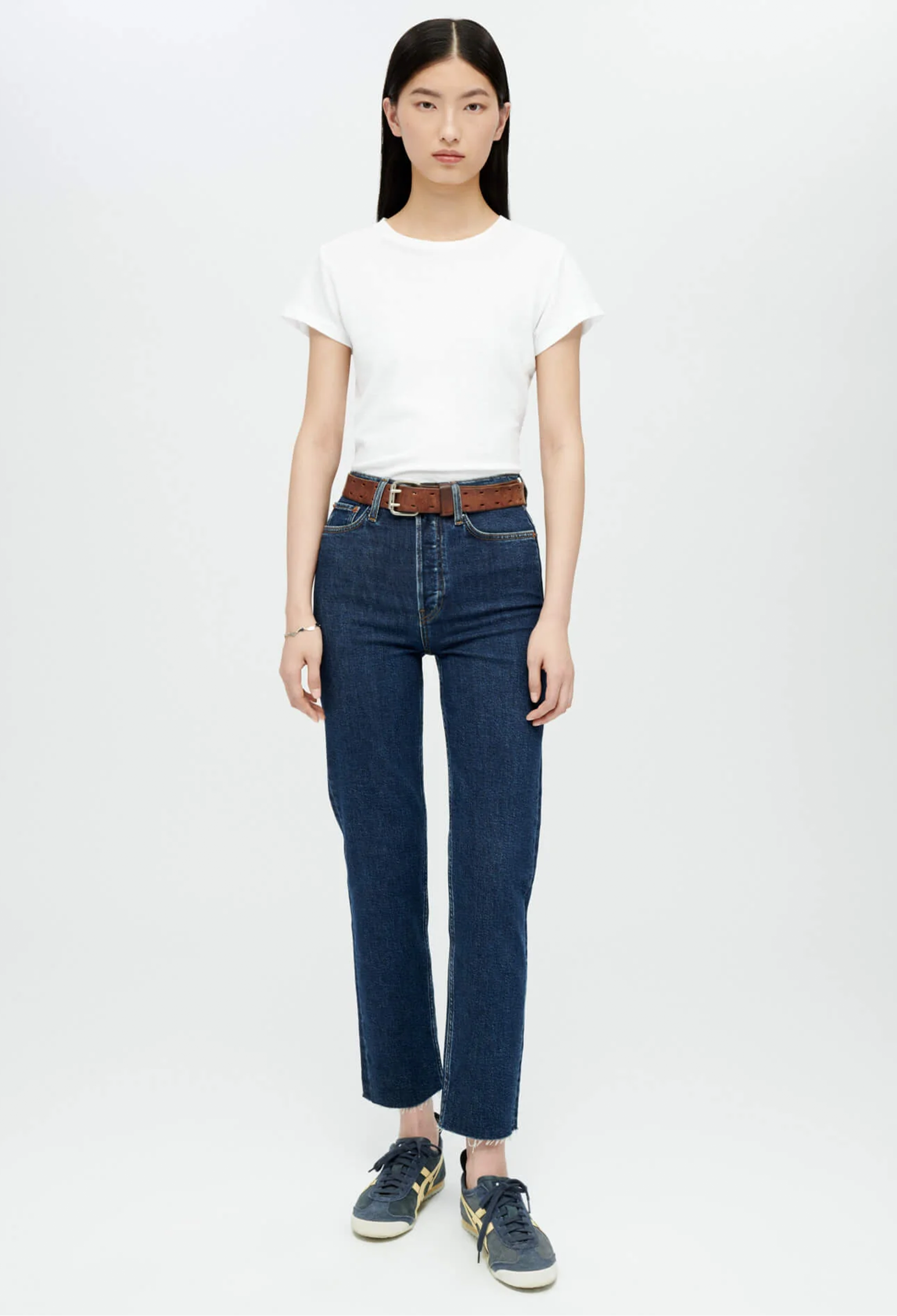 Model wears white crewneck t-shirt with dark wash blue jeans and brown belt and navy sneakers. She stands in front of a white background. 