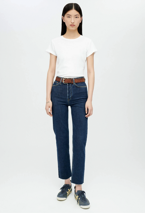 Model wears white crewneck t-shirt with dark wash blue jeans and brown belt and navy sneakers. She stands in front of a white background. 