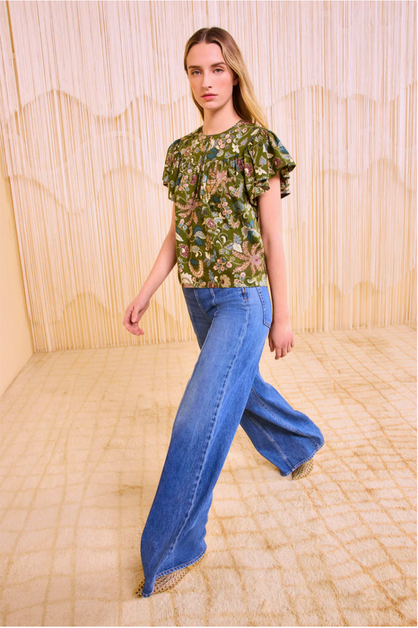 Model in the flutter sleeve green floral print blouse and high waisted light wash jeans. She is the middle of taking a step on a carpet. 