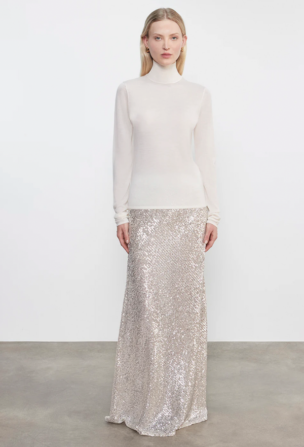Woman wearing a white turtleneck sweater and silver sequin skirt on a plain background