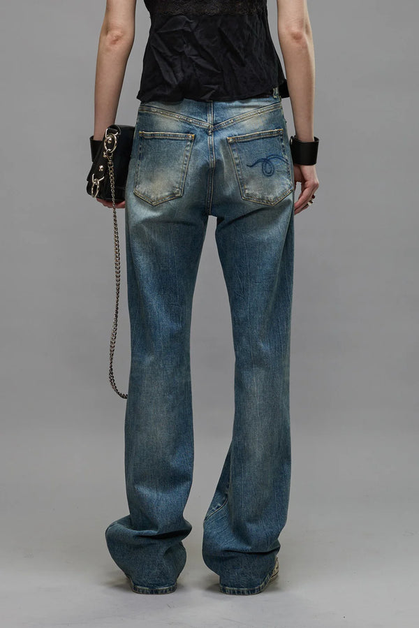 The back of a woman wearing a pair of high-rise slim blue jeans with a black belt and black top holding a brown purse. Grey background.