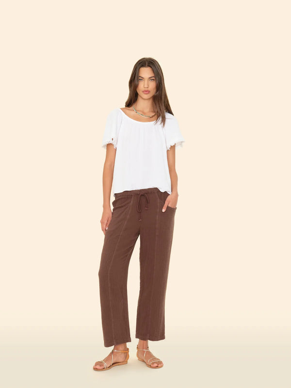 model standing in front of a slightly tinted yellow wall. she is wearing a white t-shirt and brown gauze drawstring pants. Her shoes are a gold sandal and her hand is in her pocket. 