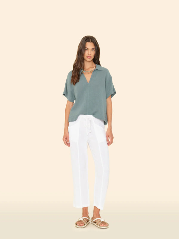 model standing in front of a slightly tinted yellow wall. she is wearing a sage green top that is a v-neck shape with a collar and white gauze drawstring pants. Her shoes are a white & taupe sandal.