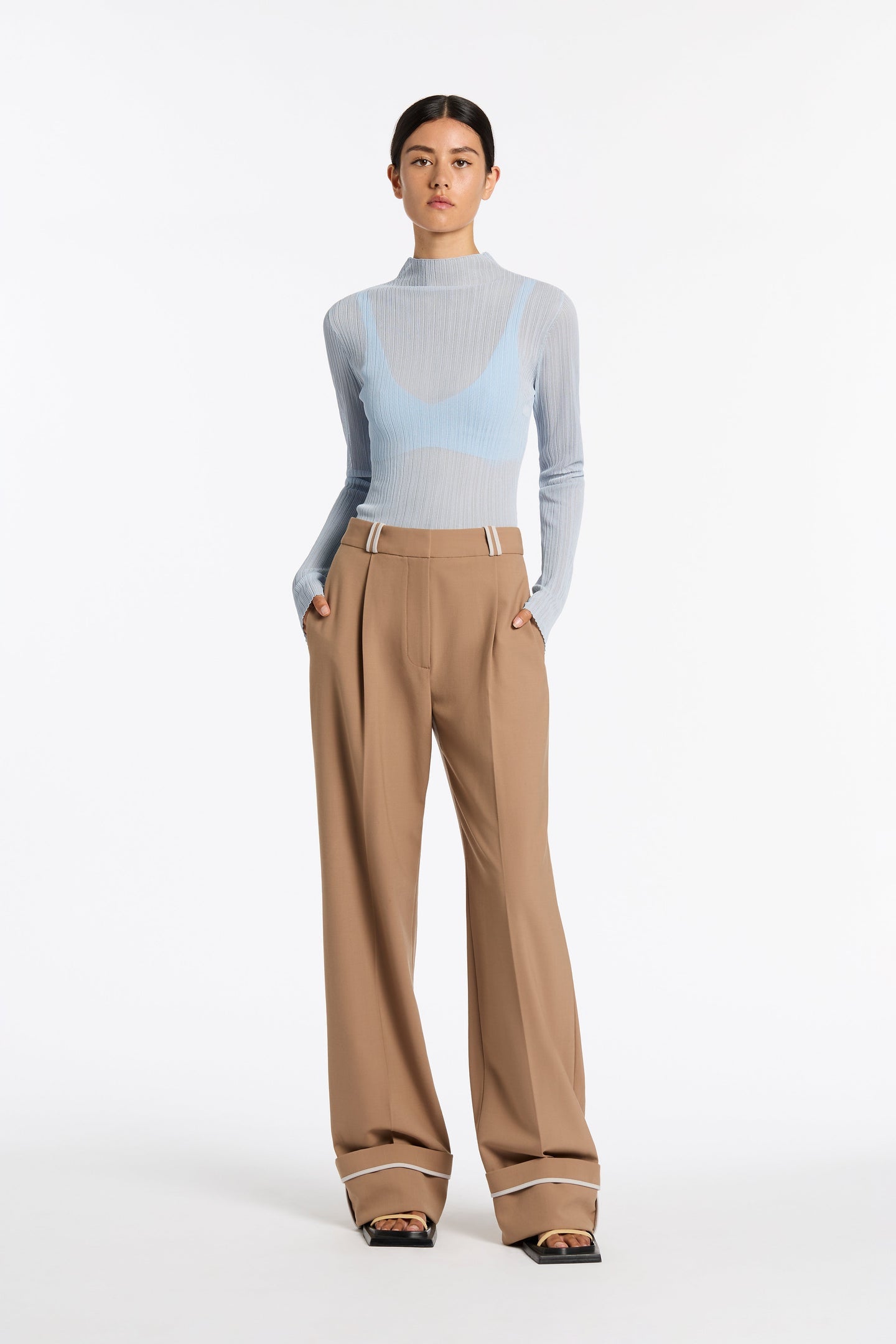 Model wears tan pants with white belt loops, black sandals with a yellow strap, and a light blue long sleeved mock neck. Set against a white background. 